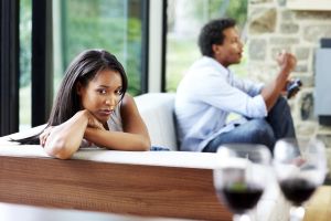 Unhappy young woman sitting thoughtfully on sofa with her husband enjoying video games in the background