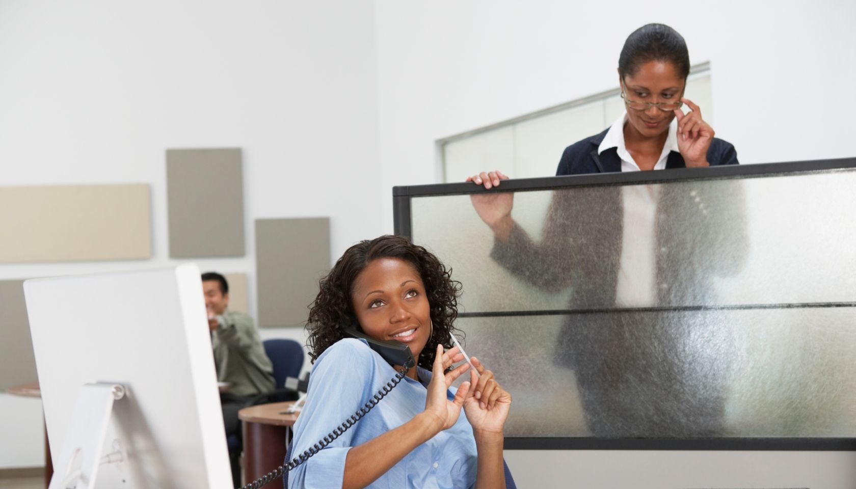 African businesswoman filing fingernails at desk with boss looking over cubicle wall