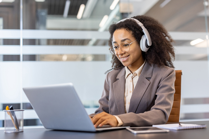 A smiling young African American woman in a suit and headphones is sitting at a desk in the office and working on a laptop