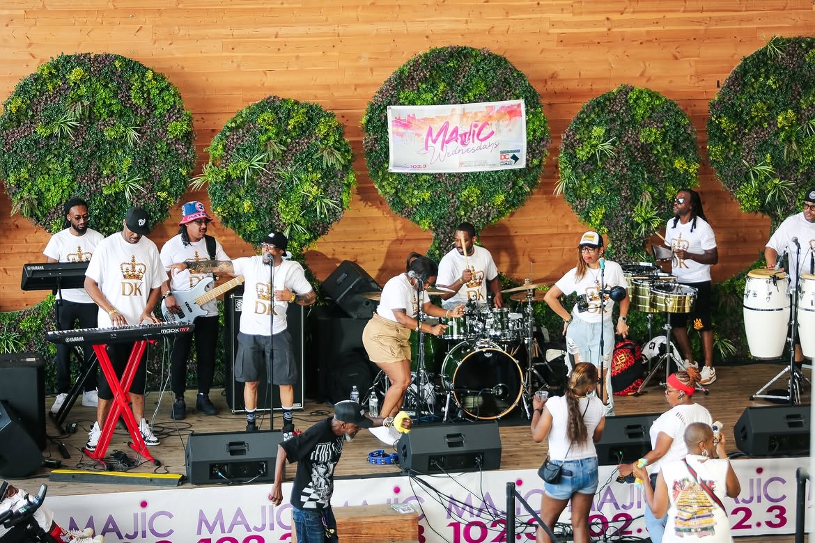 A live music performance on an outdoor stage with a band playing instruments and a crowd in the foreground. The stage is decorated with large circular moss-covered structures and a "Magic" logo.