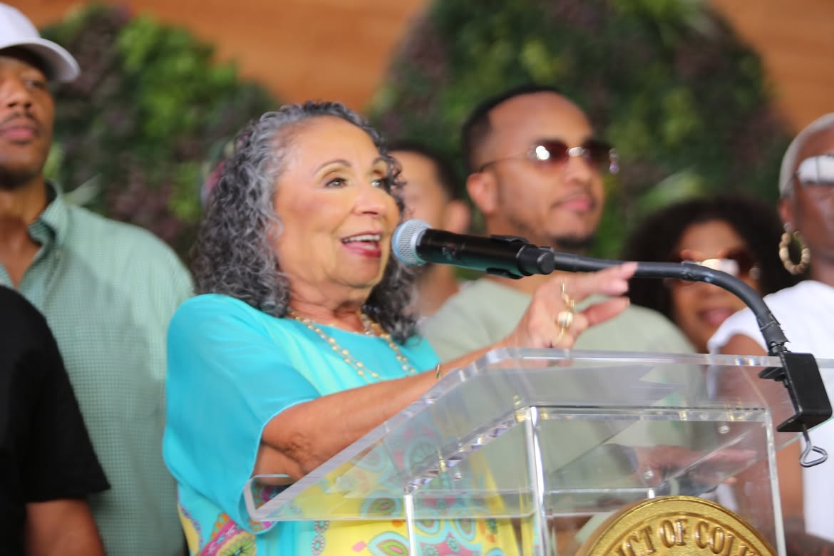 An older woman with curly gray hair speaking into a microphone at a podium, surrounded by a crowd of people.