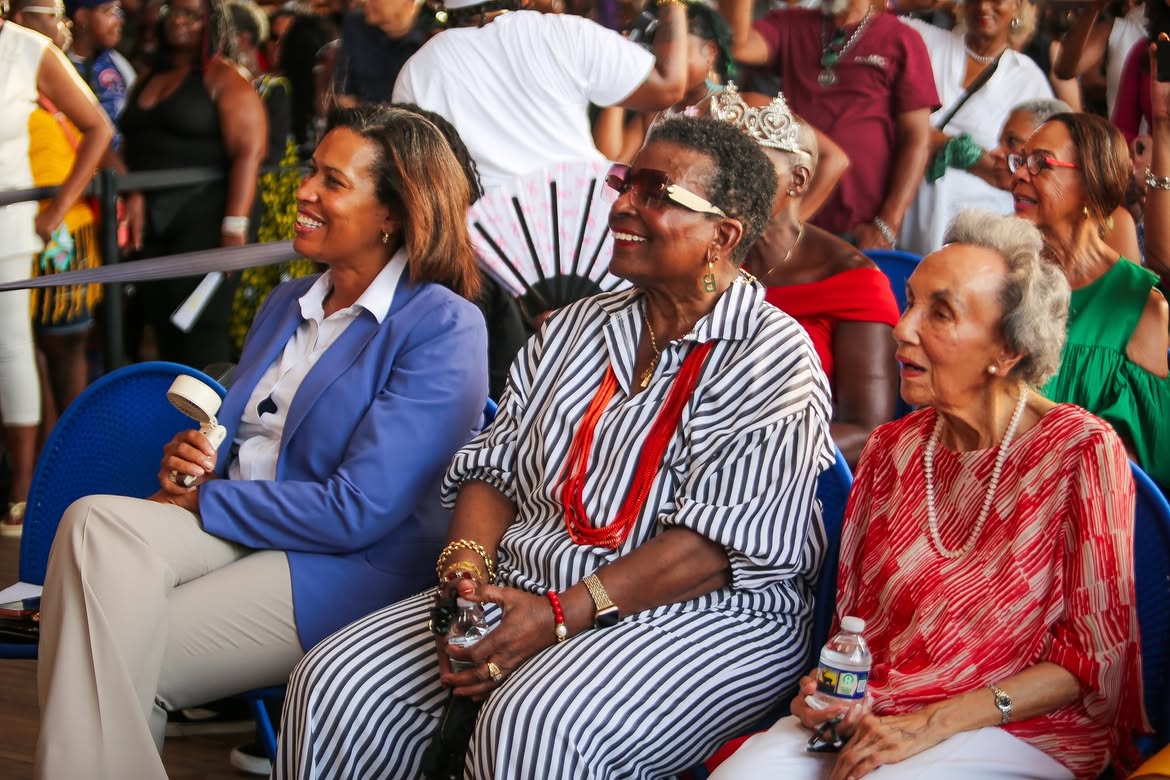 A crowd of people at an event, including several women wearing colorful, patterned clothing and accessories. One woman is wearing a crown-like headpiece.