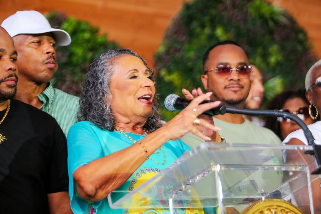 A group of people, including a woman with curly gray hair speaking into a microphone, gathered outdoors in a setting with greenery.