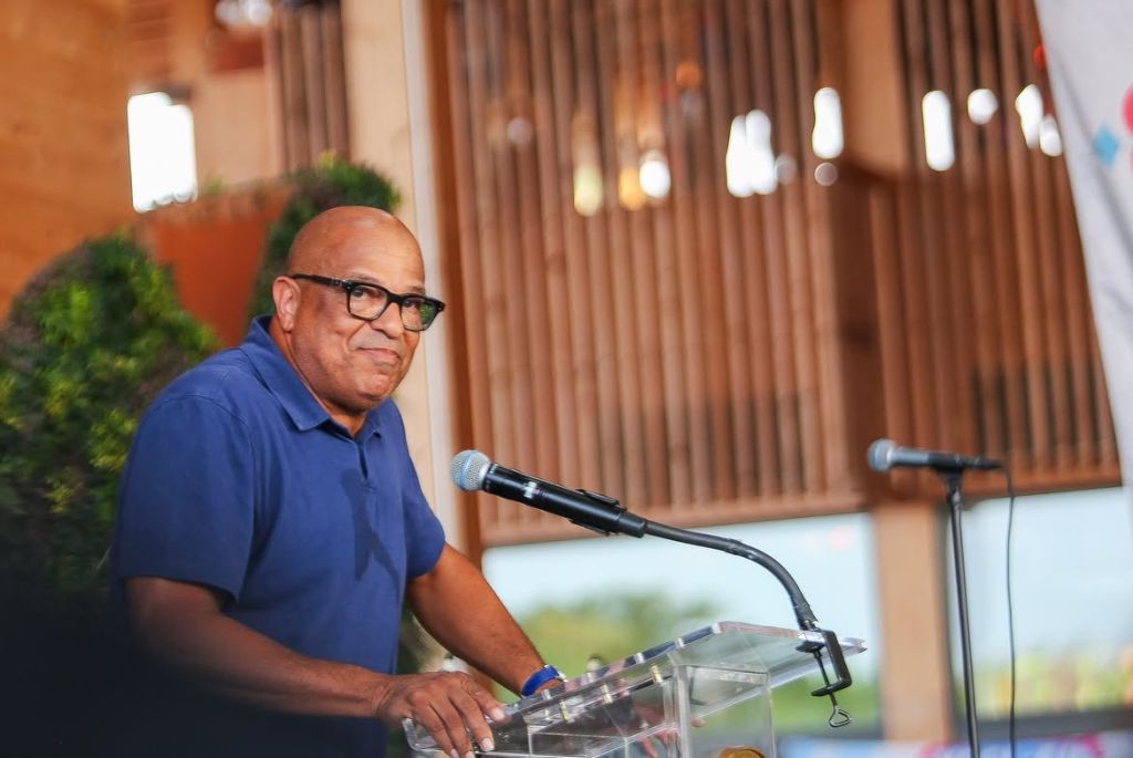 An older man with glasses speaking into a microphone at a podium, with a wooden wall backdrop and greenery visible.