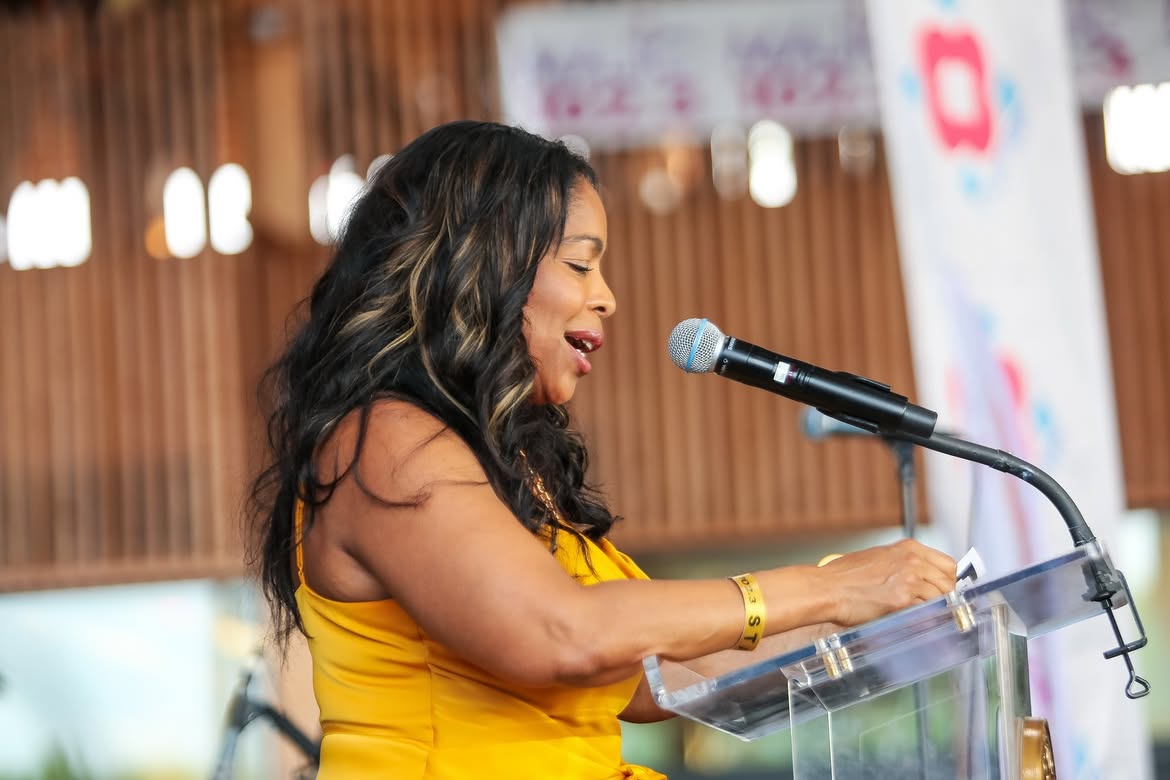 A woman in a yellow dress singing into a microphone on a stage with colorful banners in the background.