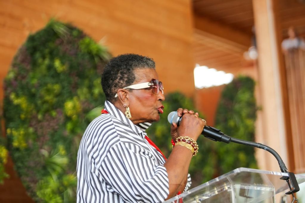 A woman wearing a striped jacket and sunglasses speaking into a microphone at a podium.