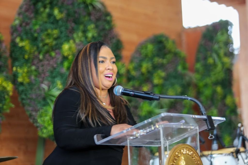 A woman speaking into a microphone at a podium, with lush greenery in the background.