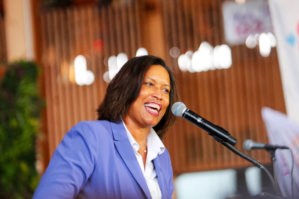 A smiling woman speaking into a microphone on a stage with a wooden backdrop and lighting.