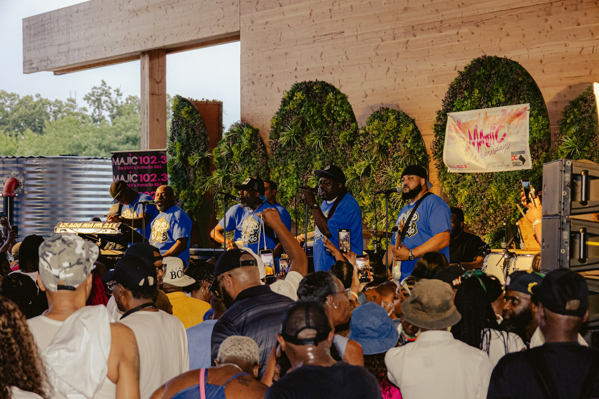 A crowd of people gathered in front of a stage with a band performing. The stage is decorated with moss-covered sculptures. The band members are wearing blue shirts with logos.