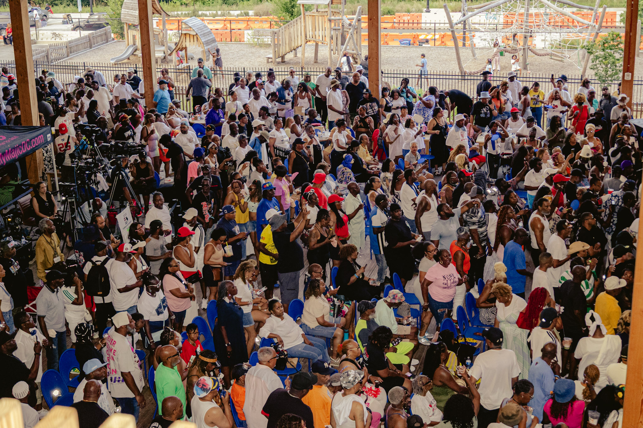 Crowded outdoor event with many people gathered, some wearing colorful clothing and holding signs or banners.