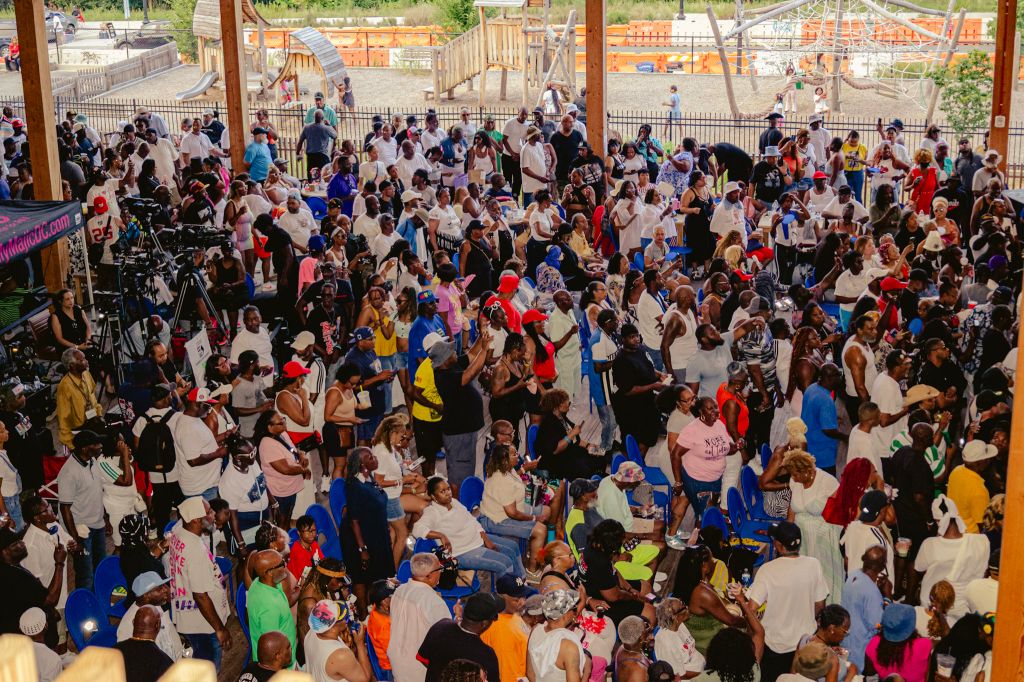 Crowded outdoor event with many people gathered, some wearing colorful clothing and holding signs or banners.