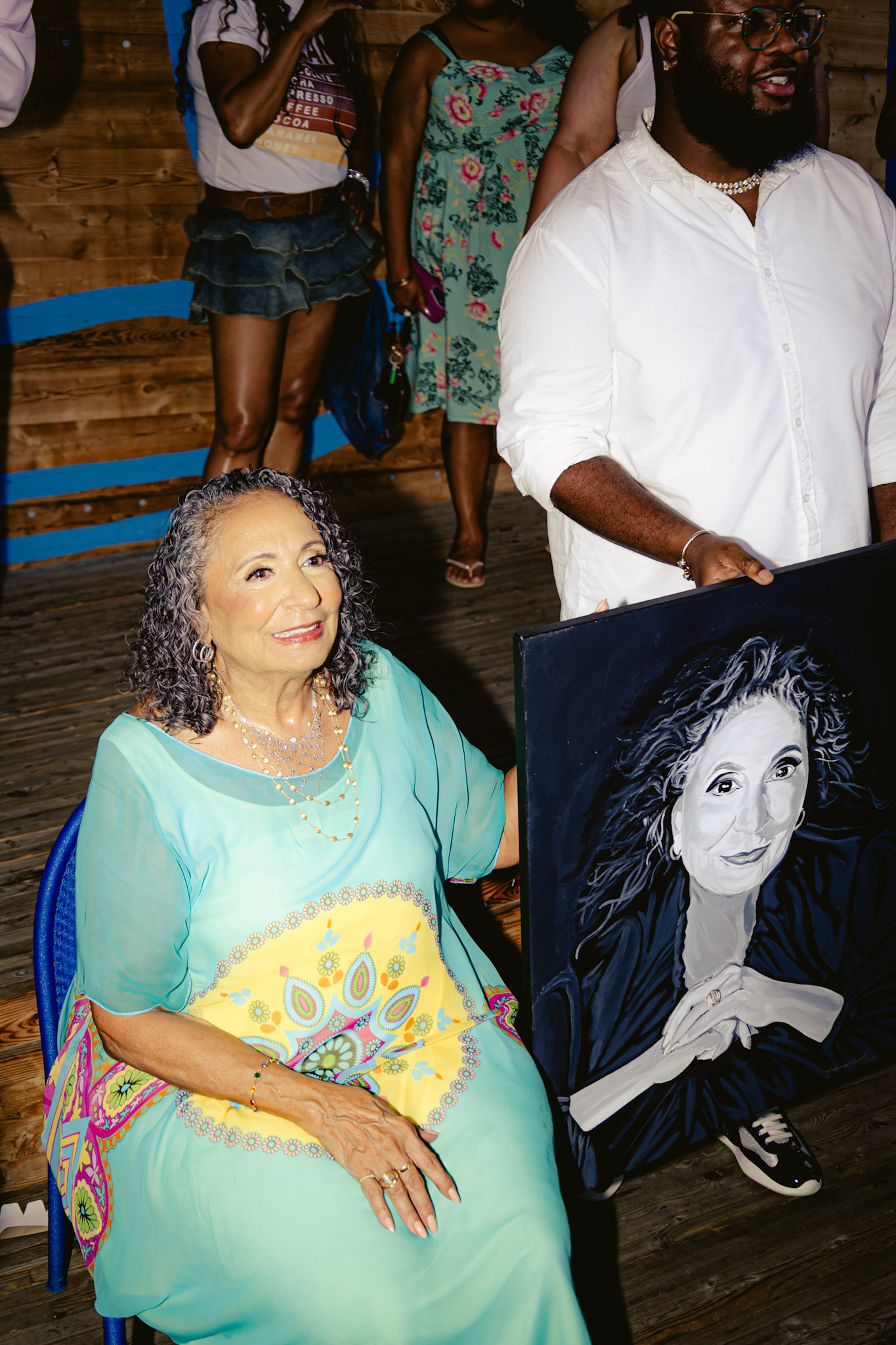 A smiling older woman in a colorful dress holding a painting of a woman's portrait, surrounded by other people in a casual setting.