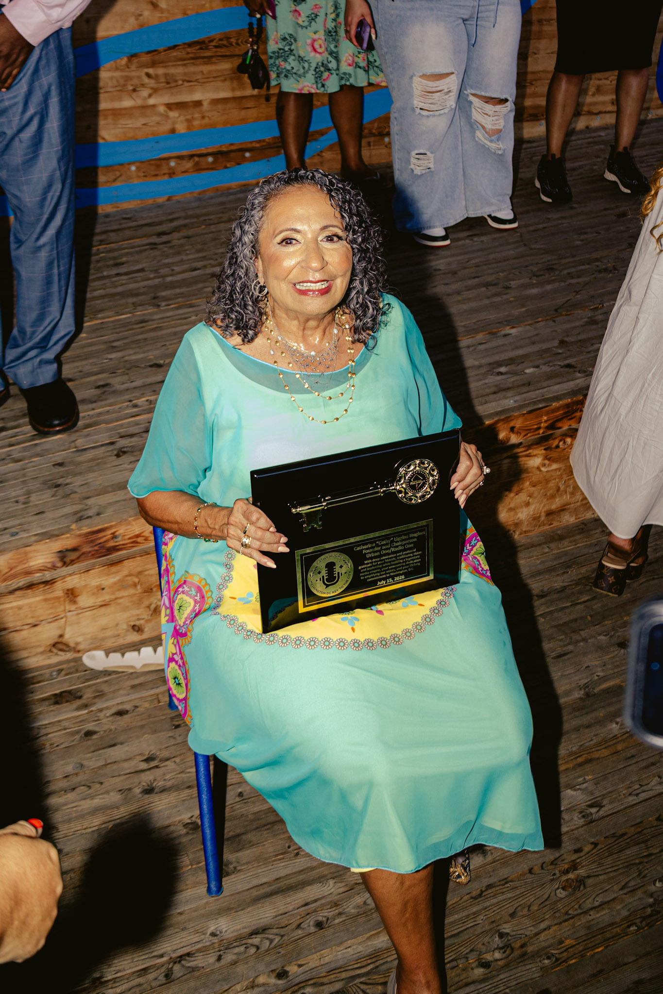 A smiling older woman in a turquoise dress holding a framed award on a wooden stage, surrounded by other people in casual clothing.