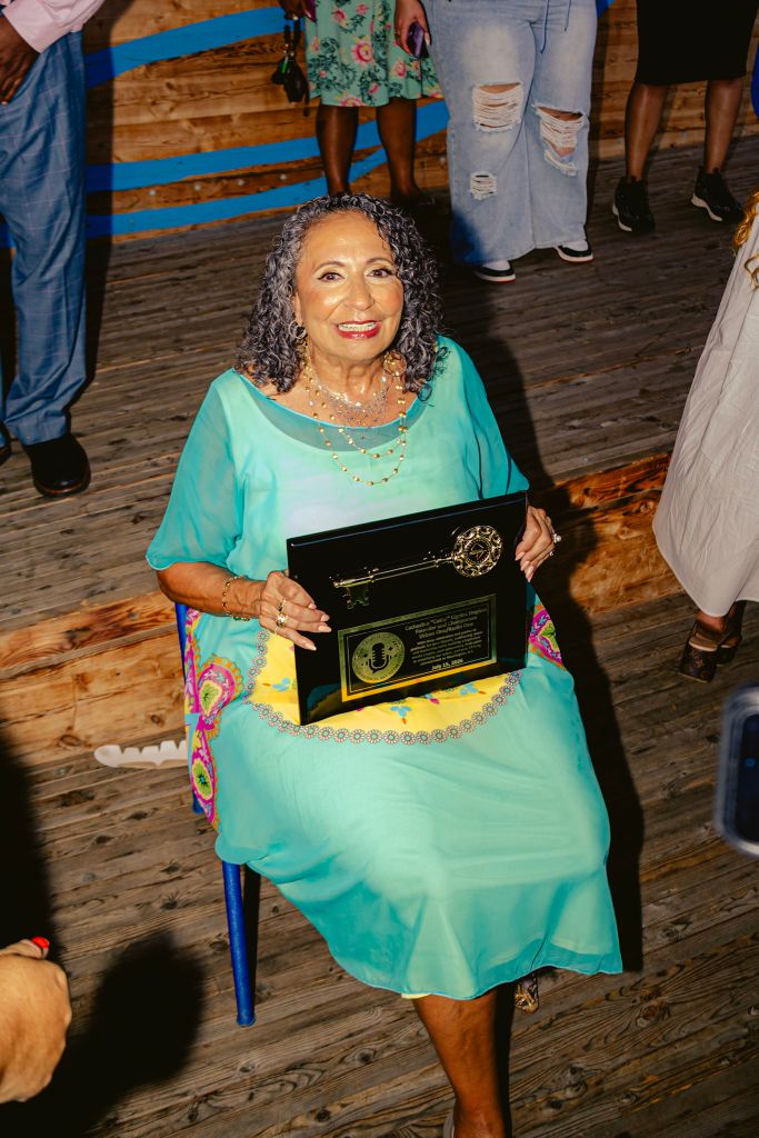 A smiling older woman in a turquoise dress holding a framed award on a wooden stage, surrounded by other people in casual clothing.