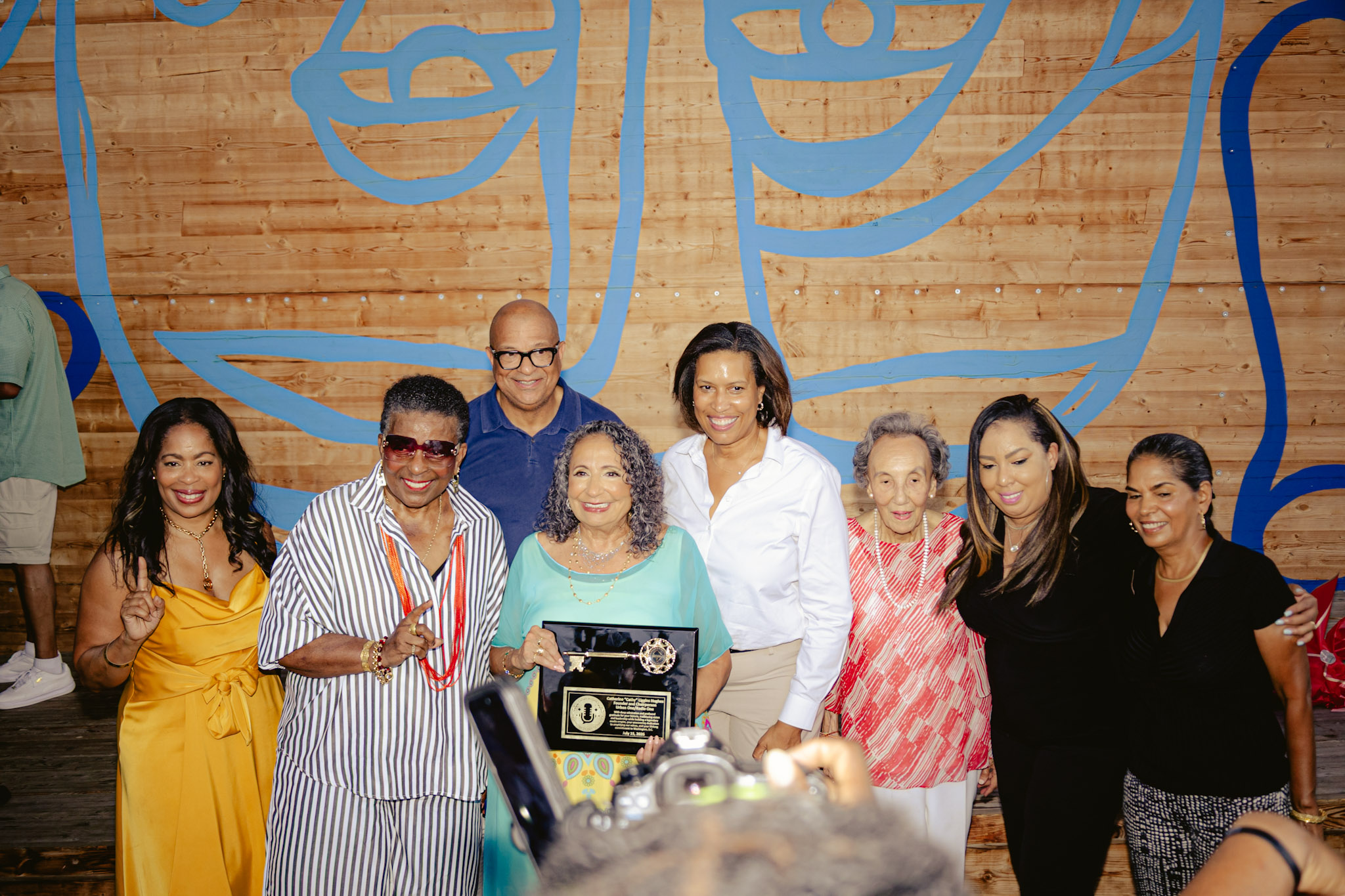A group of diverse people posing together, holding an award or recognition. The background features colorful abstract artwork on a wooden wall.