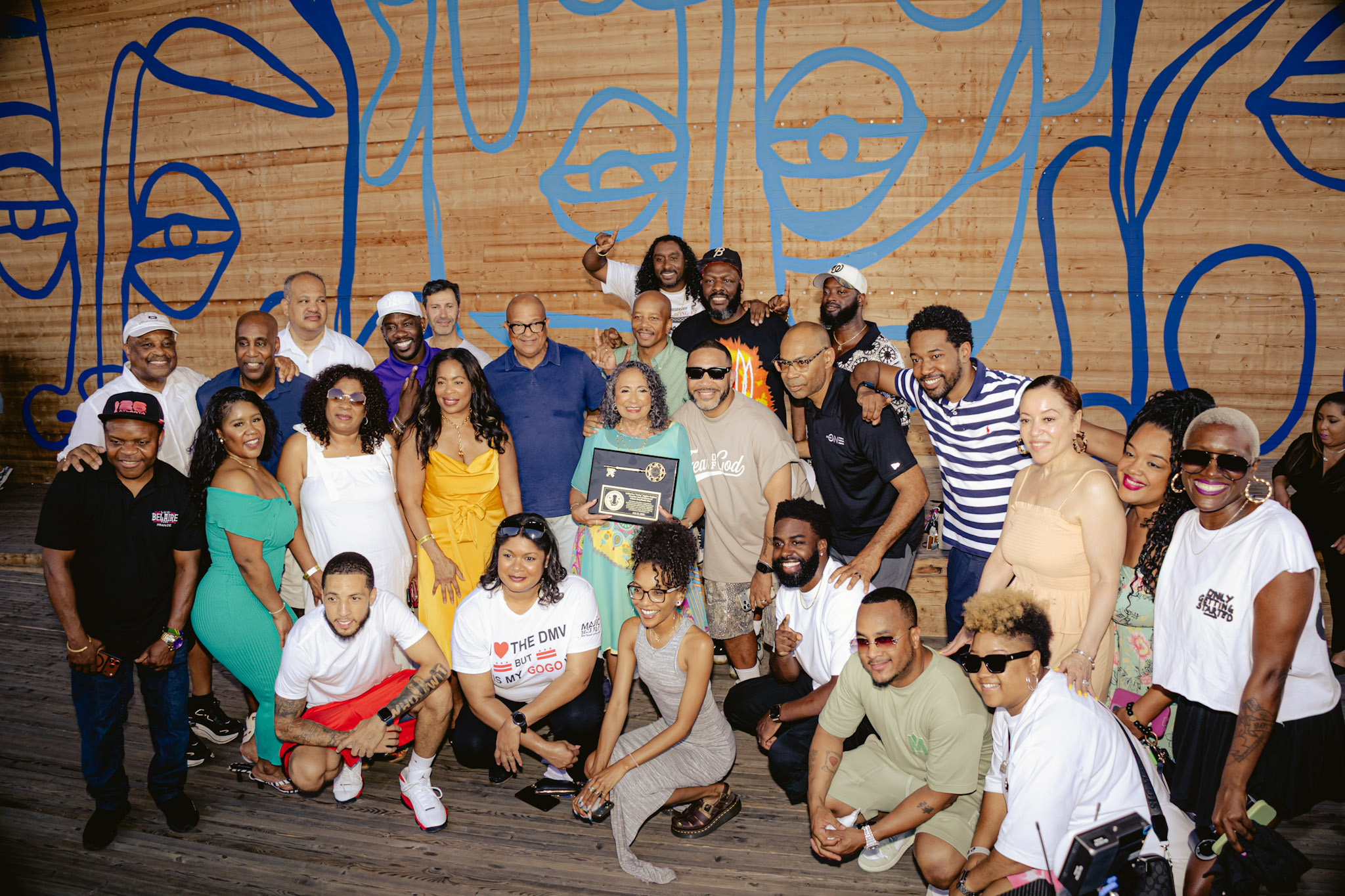 A large group of people posing together in front of a colorful mural, holding a framed award or certificate.