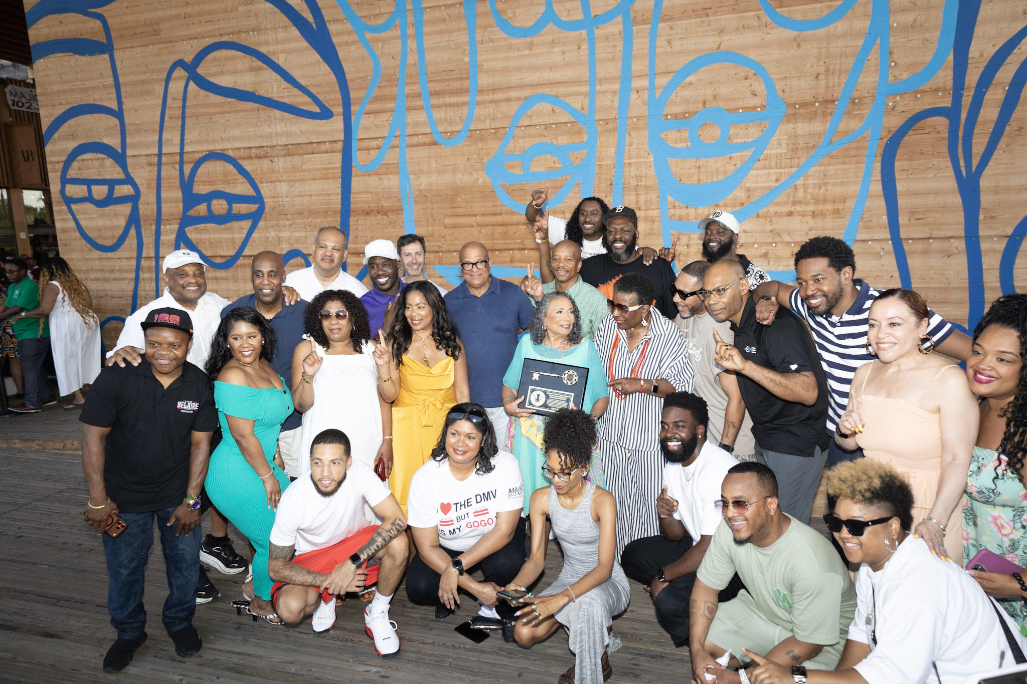 A large group of people, mostly Black, gathered in front of a colorful mural. They are celebrating and holding up a plaque or award.