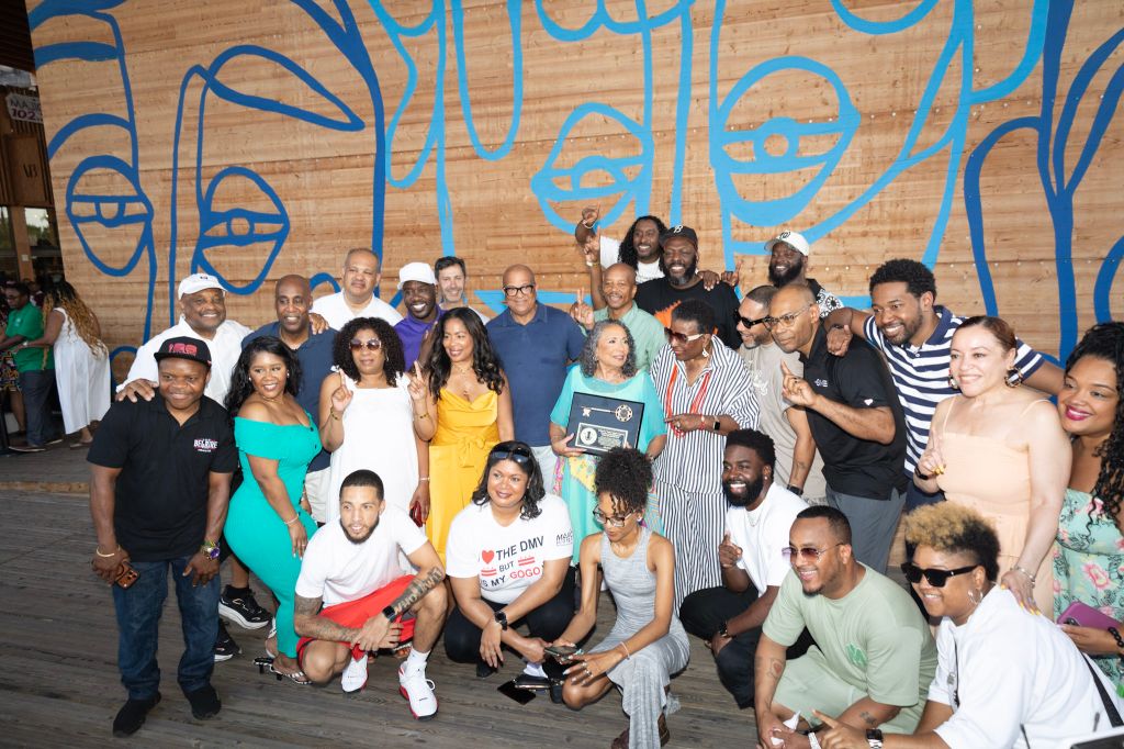 A large group of people, mostly Black, gathered in front of a colorful mural. They are celebrating and holding up a plaque or award.