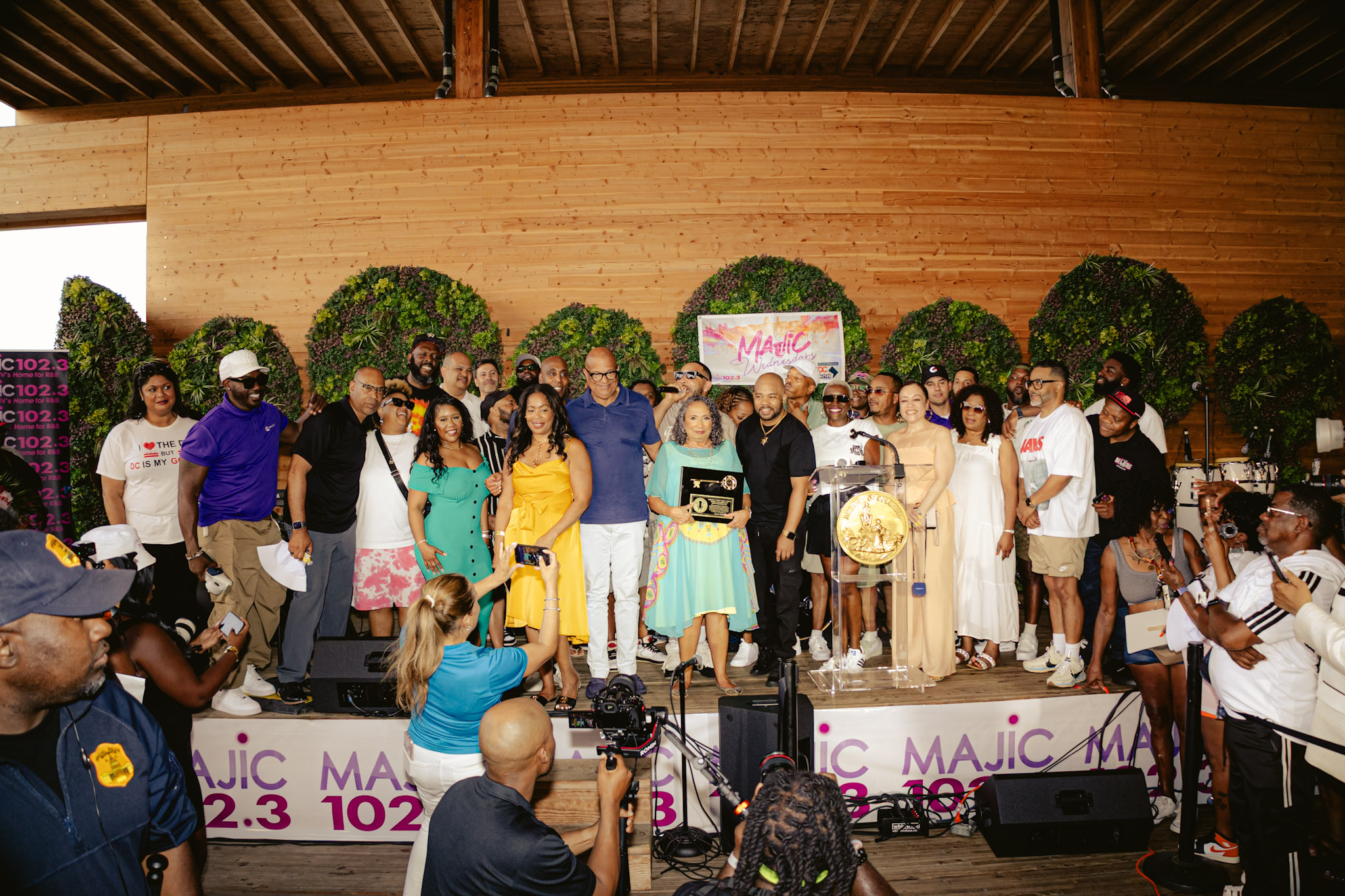 A large group of people gathered on a stage, some holding microphones and awards. The stage is decorated with green topiary shapes and the backdrop includes radio station call letters and frequencies.