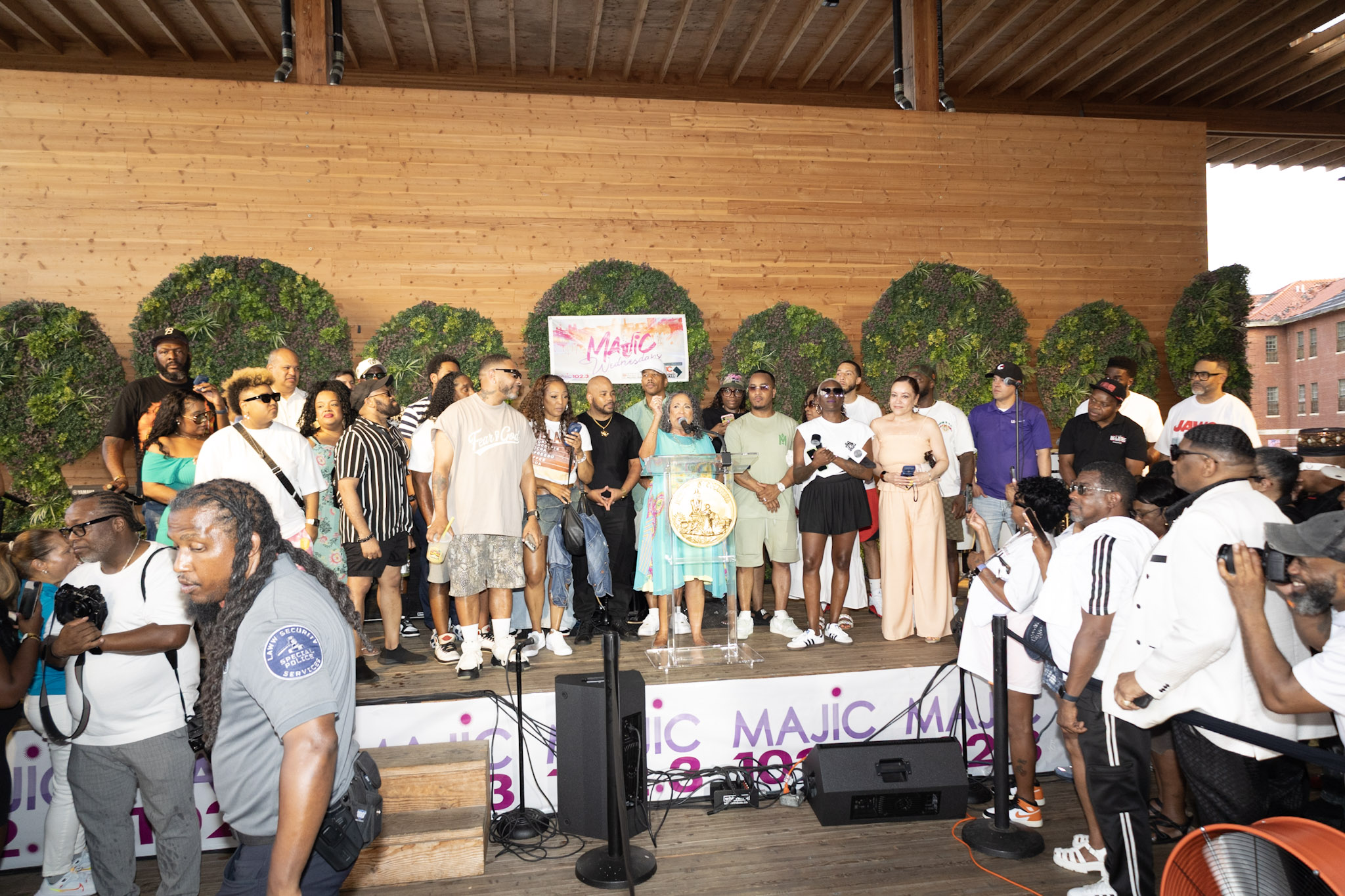 A large group of people gathered on a wooden stage, with a "Majic" logo and speakers visible. The crowd appears to be celebrating or attending an event.