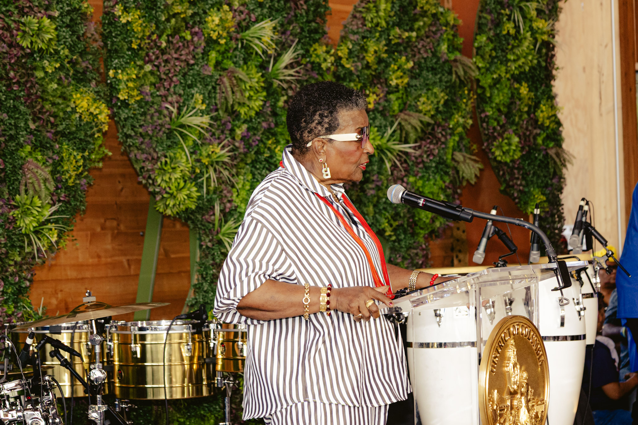 A person playing drums and percussion instruments on a stage surrounded by lush greenery.