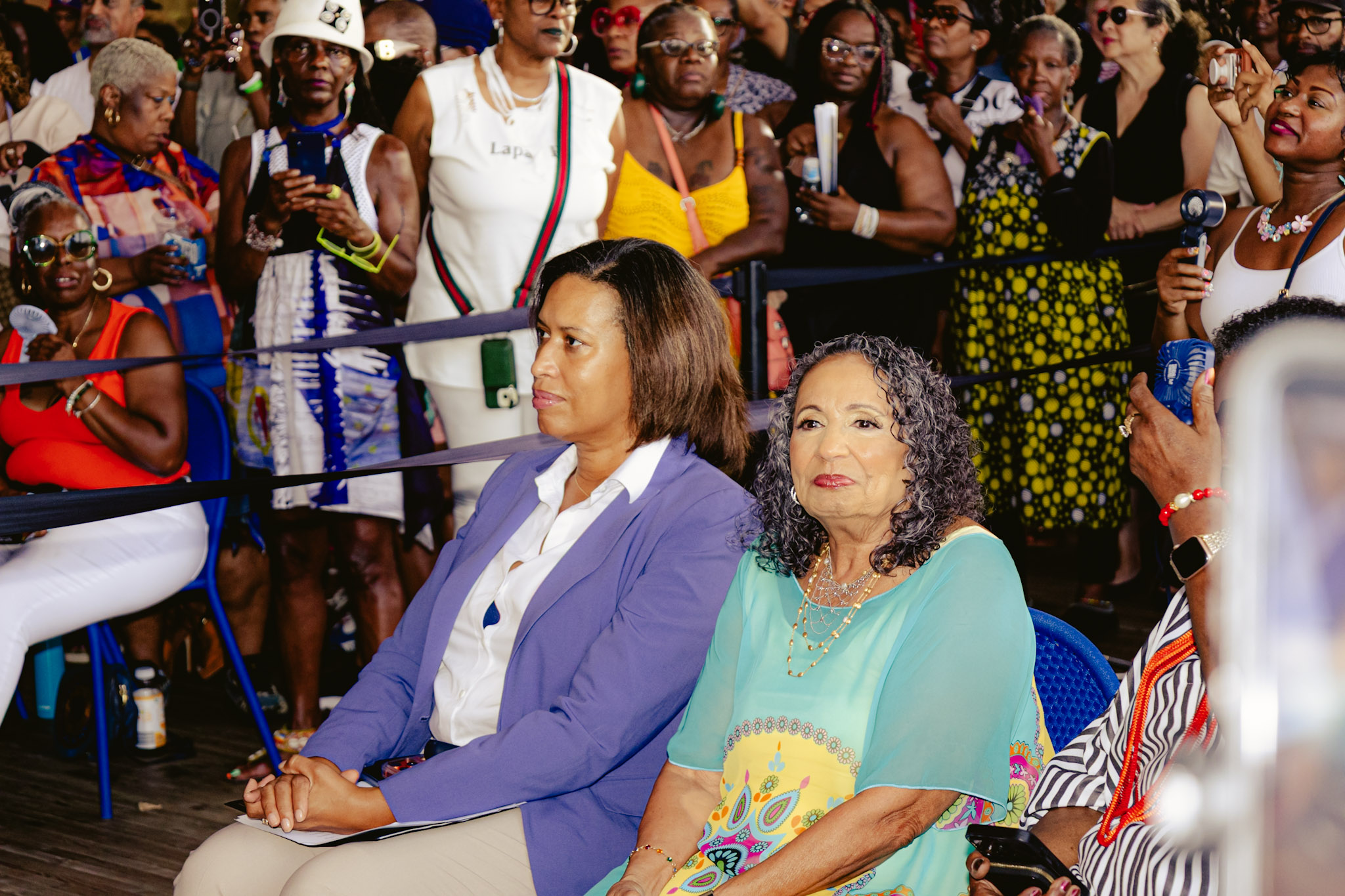 A crowd of people, many wearing colorful and festive attire, gathered at an outdoor event. Two women, one in a purple jacket and the other in a turquoise dress, are seated in the foreground.