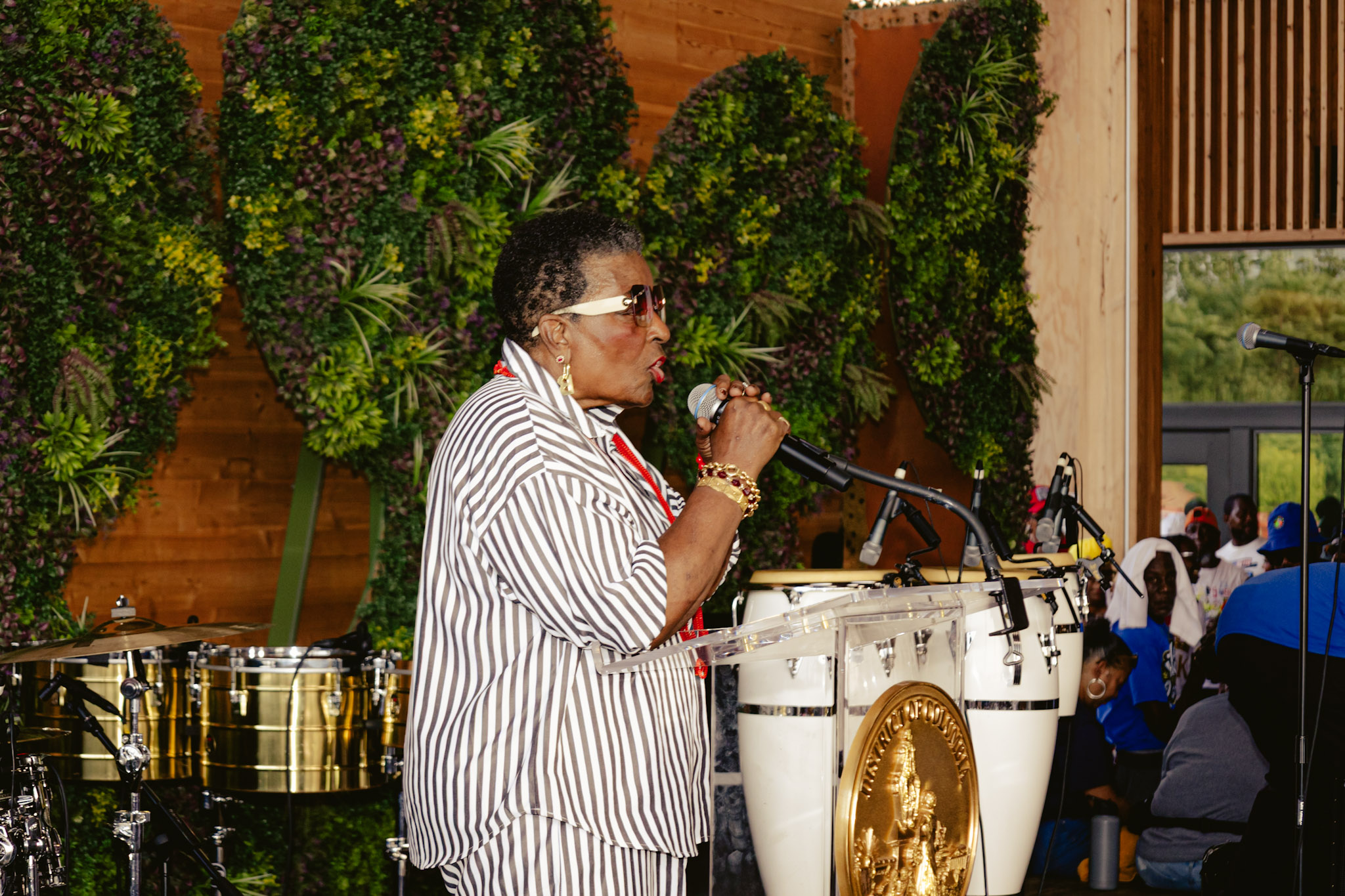 A woman in a striped jacket singing into a microphone on a stage surrounded by lush greenery.