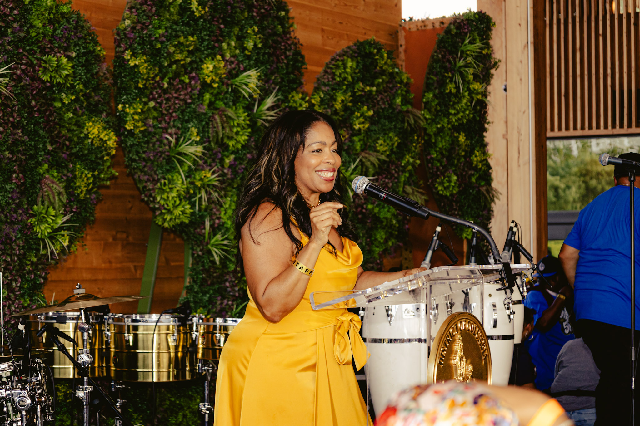 A smiling woman in a yellow dress performing on stage in front of a lush green backdrop.