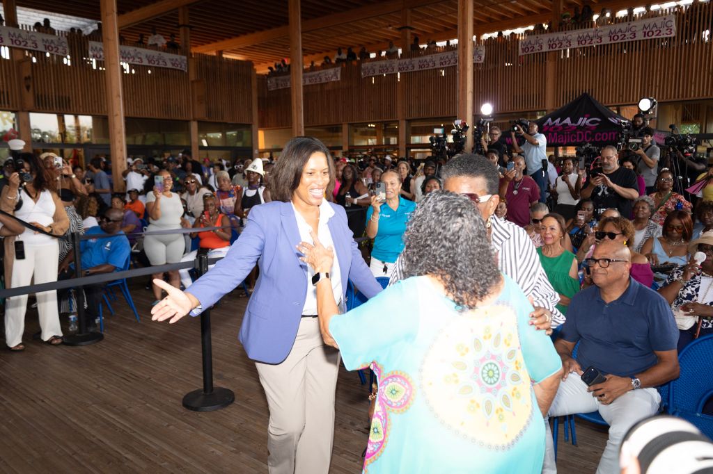 A crowded indoor event with a woman in a purple blazer greeting and interacting with a group of people. The event appears to have a "MAJIC" branding visible.
