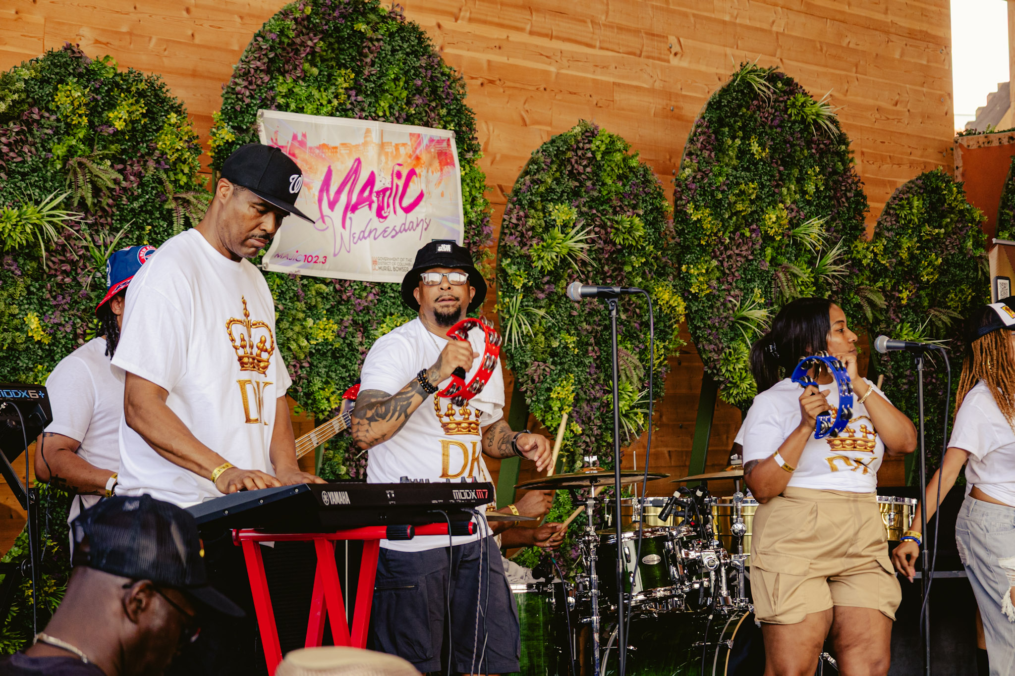 A group of musicians performing on stage in front of a backdrop with the text "Magic Wednesday" and lush greenery.