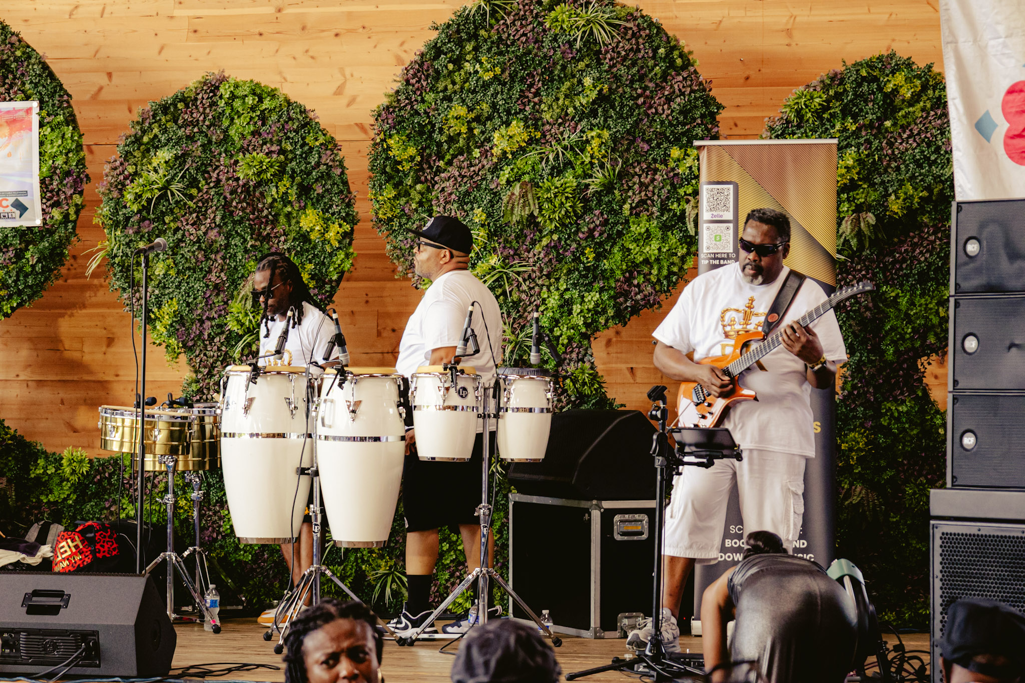 A live music performance on a stage with a lush green plant wall backdrop. The band consists of three musicians playing congas, guitar, and other instruments.