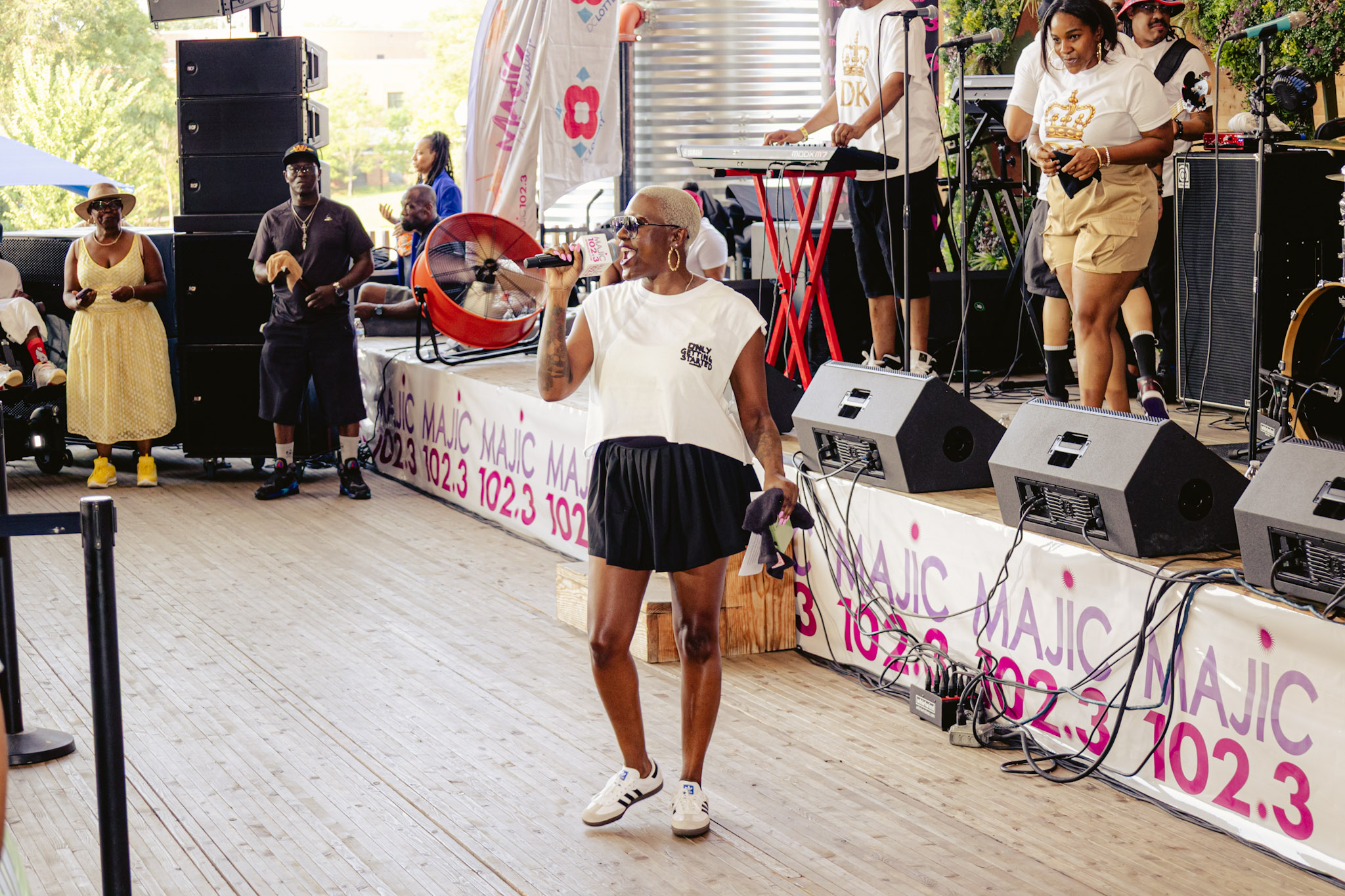 Outdoor music festival with performers on stage, speakers, and crowd in the background. Text on banners reads "MAGIC 102.3".