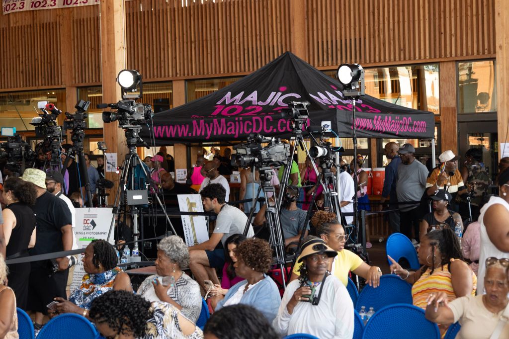 A crowded outdoor event with a large black tent displaying the "My Majic DC" logo and website. Several people are gathered around the tent, which is surrounded by cameras and other media equipment.