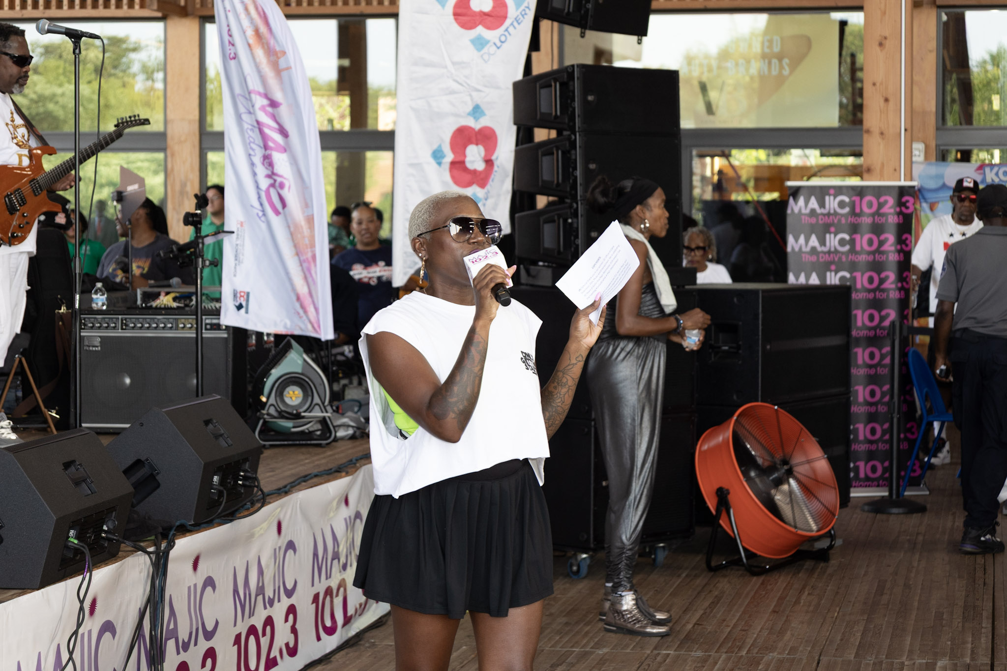 A person wearing sunglasses and a white t-shirt holding a microphone on a stage with speakers, flags, and signage for "Magic 102.3" radio station.