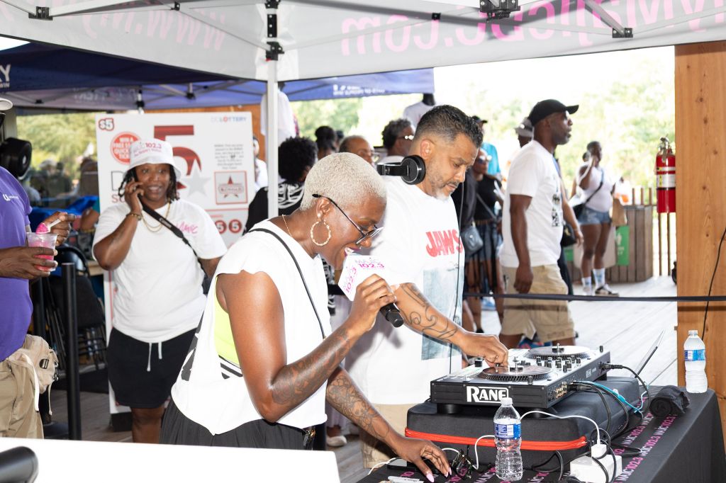 A group of people gathered at an event, with a DJ mixing music on a Rane sound system and a large screen displaying the event's branding.
